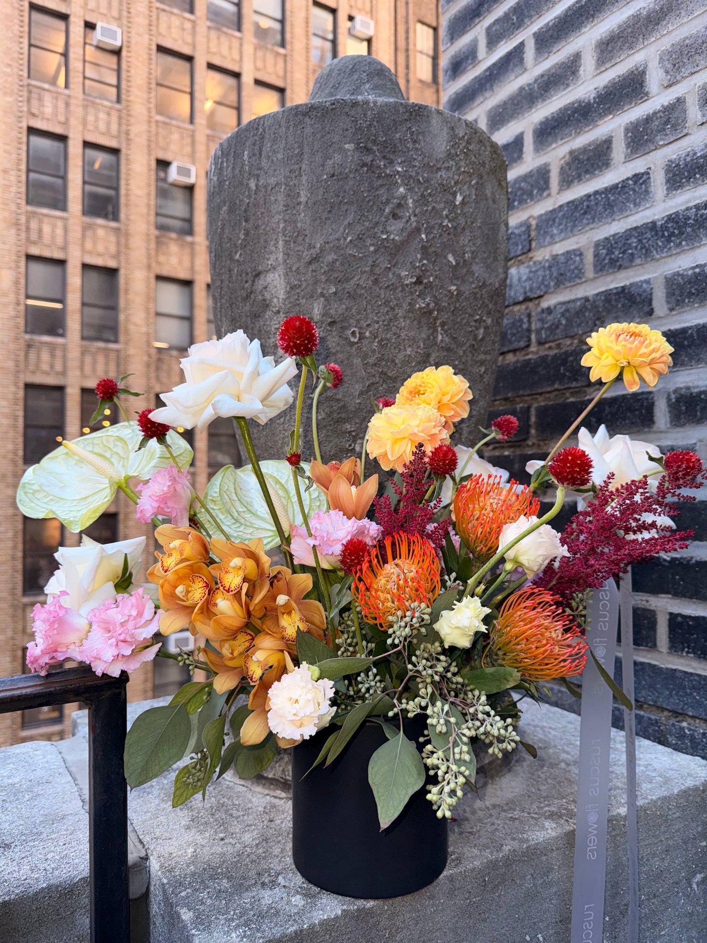 Bouquet of flowers in a black vase against a brick wall with a building in the background