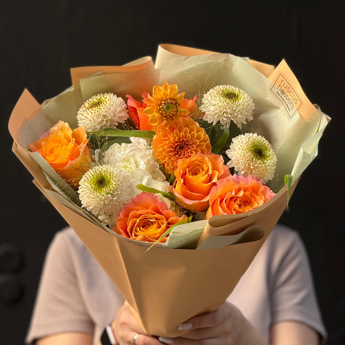 Bouquet of orange and white flowers wrapped in brown paper held by a person against a dark background